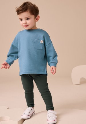 Lille dreng står iført blå langærmet skjorte, mørkegrønne leggings og hvide sneakers mod en beige baggrund med abstrakte former.