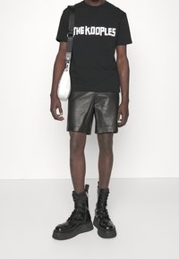 Svart t-shirt med "THE KOOPLES" grafik, lädershorts, svarta lackade snörstövlar och en vit axelväska. Släta texturer, modern design.