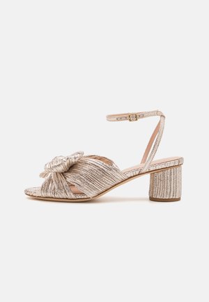 Strappy heeled sandal in metallic light gold with a knotted design, open toe, and a block heel. Features textured fabric and adjustable ankle strap.