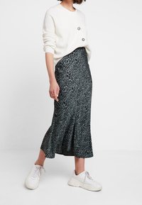 Suéter blanco acanalado con dos botones decorativos, combinado con una falda midi de estampado de leopardo verde. La modelo lleva zapatillas blancas.