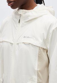 Κλείσιμο πλάνου ενός ατόμου που φοράει ανοιχτό μπεζ ζακέτα με κουκούλα και φερμουάρ Columbia, με ορατό λογότυπο στο στήθος.