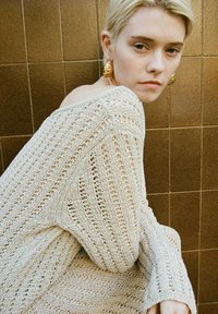 Häkelndes cremefarbenes Sweatshirt mit weiten Ärmeln und offenem Webmuster, das eine weiche Textur aufweist. Goldene Statement-Ohrringe vervollständigen den Look.
