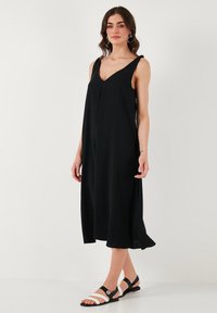 Čierne šaty bez rukávov s výstrihom do V, voľná silueta a mäkká látka; nosené s čierno-bielymi sandálmi. Jednoduchý, elegantný dizajn.