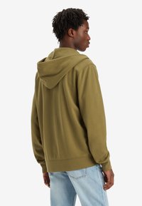Oliven grønn hettegenser med hette, laget av mykt materiale. Har en avslappet passform og ribbestrikkede mansjetter. Brukt med lyseblå jeans.