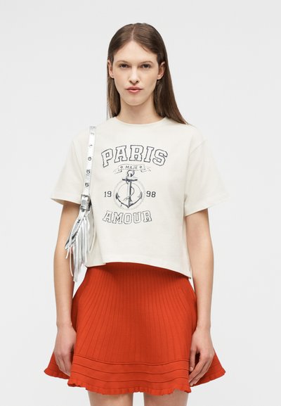 Γυναίκα που φοράει λευκό κοντό μπλουζάκι με το σχέδιο "Paris Amour", κόκκινη πλισέ φούστα και ασημένια τσάντα ώμου με κρόσσια, σε λευκό φόντο.