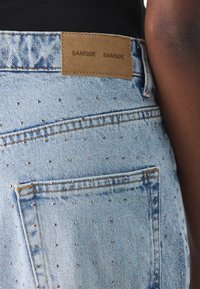 Jeans in denim azzurro con piccoli chiodi d'argento e un'etichetta in pelle marrone con la scritta "SAMSØE SAMSØE" sulla vita.
