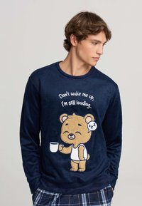 Giovane uomo che indossa un felpa blu navy con un cartone animato di un orso assonnato che tiene una tazza di caffè e il testo "Non svegliarmi, sto ancora caricando..."