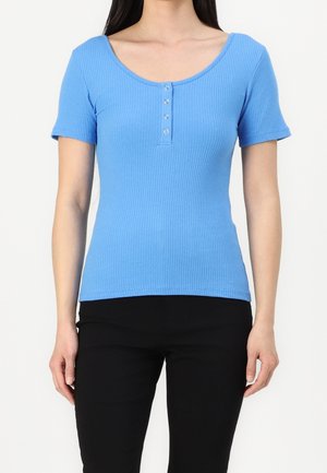 Vrouw draagt een nauwsluitend blauw geribbeld topje met korte mouwen, een ronde hals en vier zilveren knopen aan de voorkant, gecombineerd met een zwarte broek.