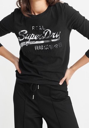 Femme portant un haut noir à manches longues avec le logo "Real SuperDry" et un pantalon noir avec cordon, les mains reposant sur les hanches.