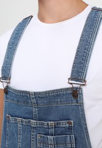 Pantalones de mezclilla con tirantes azules ajustables, herrajes en tono cobre y detalle en el bolsillo delantero. Se llevan sobre una camiseta blanca básica.