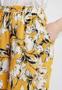 Kalhoty s květinovým potiskem s jasně žlutým pozadím, na kterém jsou bílé květy a zelené listy, vyrobené z lehké látky s elastickým pasem.