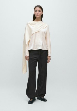 Kvinde står iført asymmetrisk cremet silketop med draperet detalje, sorte bukser med brede ben og sorte flade sko mod ensfarvet baggrund.