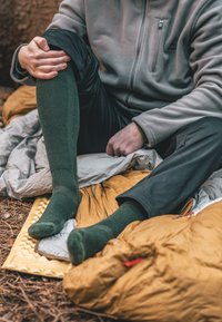 Grønne strikkede sokker båret af en person, parret med en grå fleece og sorte bukser, siddende på en gylden soveunderlag og en grå sovepose.