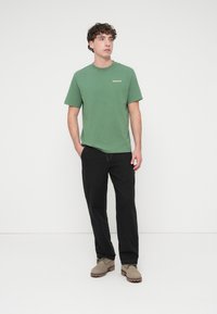Green cotton t-shirt with short sleeves and a crew neck, featuring a small logo on the chest. Paired with black pants and tan shoes.