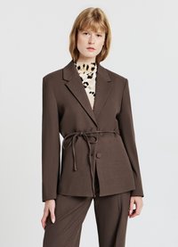 Blazer marrone con cintura, maniche lunghe, revers a punta e due bottoni frontali. Sotto, una maglietta con un motivo di dettagli animal print.