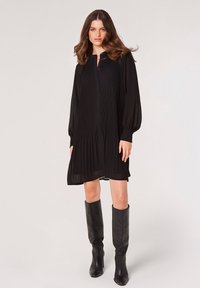 Schwarzes plissiertes Kleid mit langen Ärmeln und Kragen mit Knöpfen. Tragen mit knielangen schwarzen Lederstiefeln, die eine schlanke Silhouette schaffen.