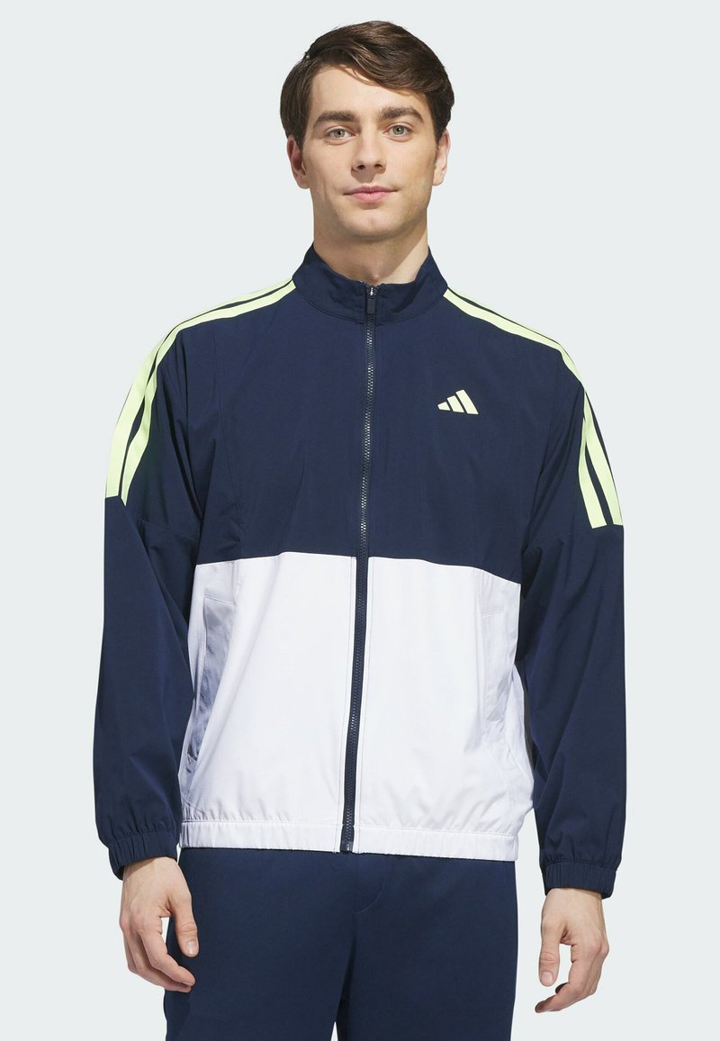 Homme portant une veste de sport zippée bleu marine et blanche avec des rayures vert clair sur les manches, debout face à l'avant sur un fond uni.