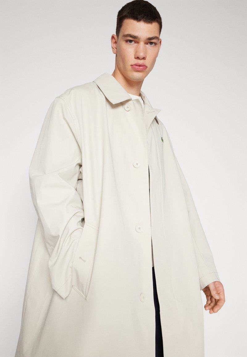 Impermeabile beige oversize con chiusura a bottoni, ampio colletto, tasche laterali e un sottile logo sul petto. Tessuto morbido al tatto.