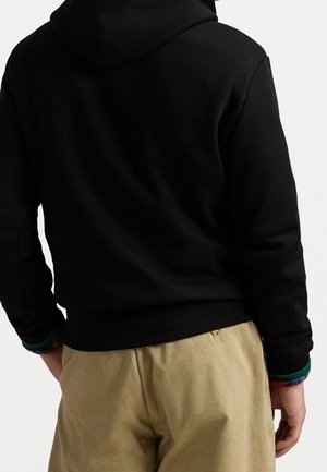 Mand iført sort hættetrøje og beige bukser, vist bagfra mod en ensartet lys baggrund.