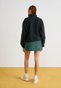 Pullover in pile verde scuro con colletto alto, abbinato a una gonna in corduroy verde chiaro. La modella indossa sneakers alte bianche e rosa.