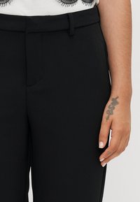 Juodos tailored kelnės su lygia tekstūra, turi diržo kilpą, šonines kišenes ir siaurą kirpimą. Ant rankos matosi gyvatės tatuiruotė.