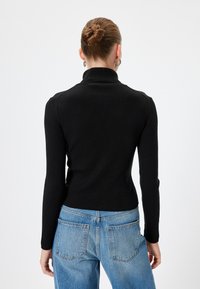 Maglione nero a costine con collo alto, maniche lunghe, taglio aderente e dettagli sullo spallone, abbinato a pantaloni a gamba larga blu chiaro.