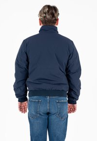 Giacca bomber blu navy con colletto alto, polsini e orlo a coste. Indossata con jeans in denim azzurri. La vista posteriore mostra la texture del tessuto liscia.