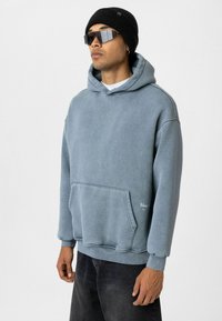 Lichtblauwe, oversized hoodie van zacht materiaal met een voorzak en geribde cuffs. Gecombineerd met een zwarte beanie en zonnebril.