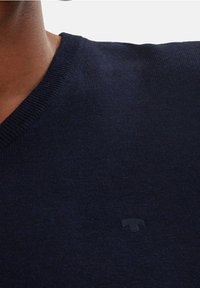 Maglione a V scollo in navy realizzato in morbido tessuto testurizzato. Presenta un sottile dettaglio del logo vicino al collo. La vista ravvicinata mette in evidenza le cuciture.