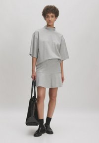 Grijze oversized shirt met een hoge kraag, gecombineerd met een bijpassende rok. Model houdt een zwarte leren tas vast en draagt zwarte sokken en schoenen.