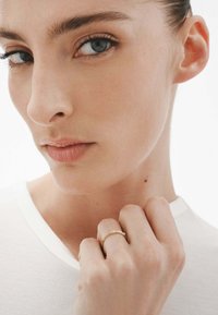 Primer plano de la cara de una persona con piel clara, ojos azules y un color de labios sutil, que lleva un anillo de oro en su dedo y una camisa blanca.