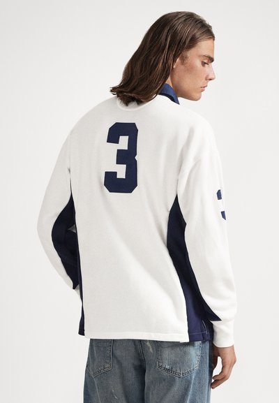 Άτομο με καστανά μαλλιά μέχρι τους ώμους, φοράει λευκό και ναυτικό μπλε μακρυμάνικο πουκάμισο με τον αριθμό 3 στην πλάτη και τζιν παντελόνι.