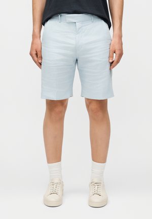 Mężczyzna w jasnoniebieskich szortach, białych skarpetkach i beżowych trampkach stojący na jednolitym tle.