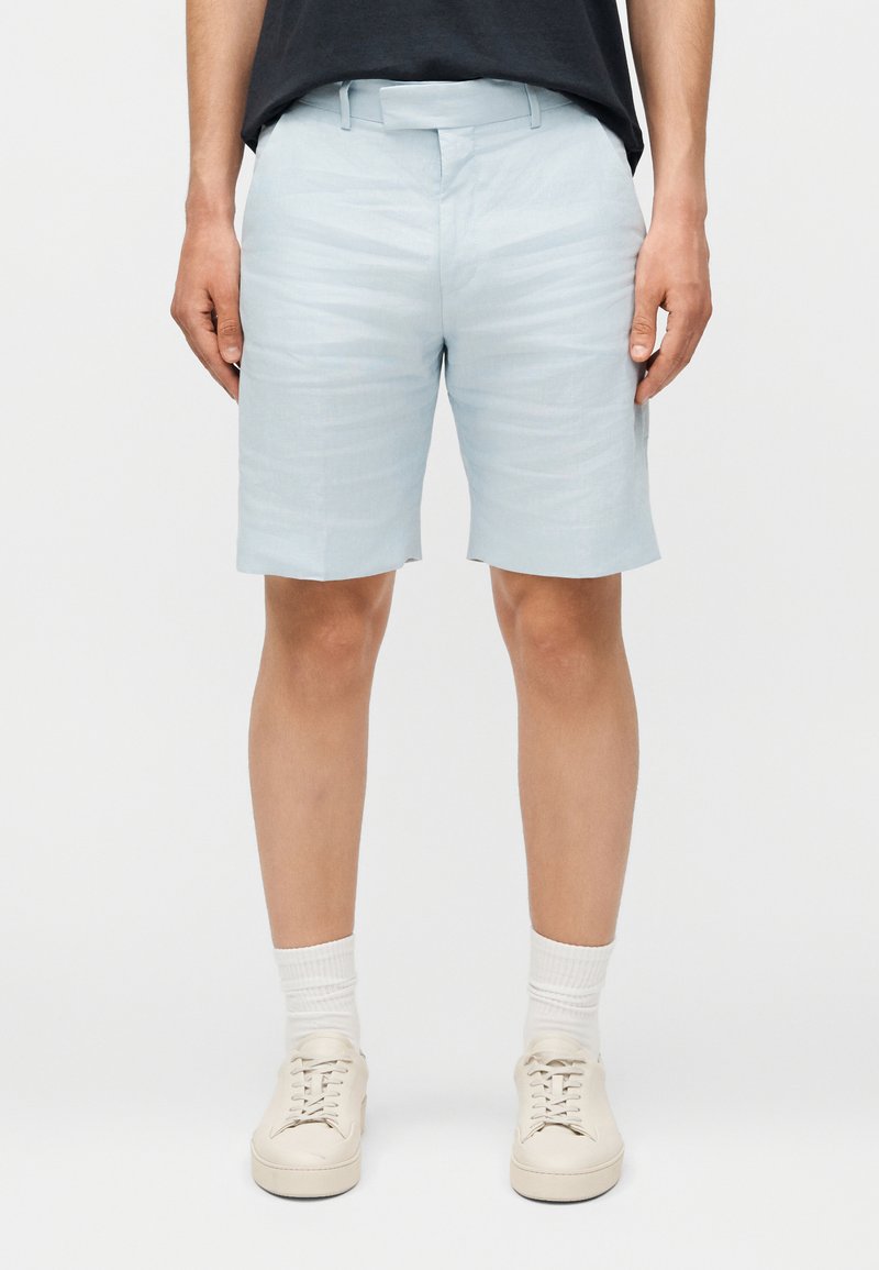 Άνδρας με ανοιχτό μπλε σορτς, λευκές κάλτσες και μπεζ αθλητικά παπούτσια στέκεται μπροστά σε απλό φόντο.