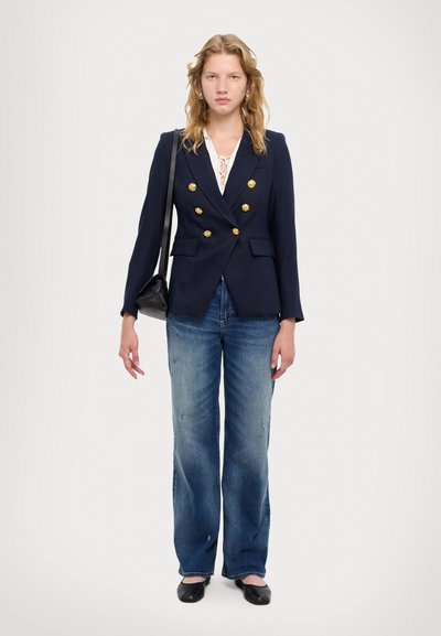 Blazer azul marino de doble botonadura con botones dorados, blusa de encaje blanca, vaqueros de pierna ancha color azul claro y bailarina negras. La modelo sostiene un bolso pequeño negro.