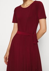 Bordeaux klänning med korta ärmar, rund halsringning och ett matchande midjeband. Tillverkad av ett mjukt tyg med en svävande design och veckade detaljer.