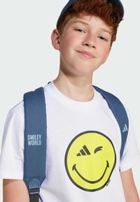 White t-shirt features a large yellow smiley face graphic. The boy wears blue padded backpack straps with "SMILEY WORLD" text.
