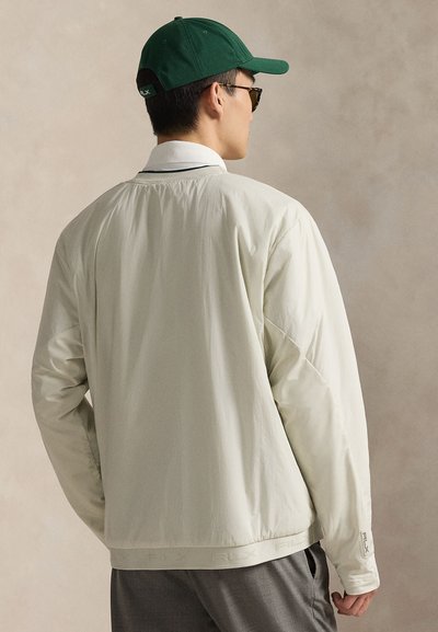 Λευκό ελαφρύ μπουφάν με μακριά μανίκια και ριμπ γιακά. Διαθέτει λογότυπο στο κάτω μέρος, φορεμένο πάνω από γκρι παντελόνια, με πράσινο καπέλο.