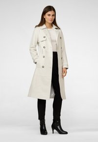 Light beige trench coat with a double-breasted design, large buttons, and side pockets, paired with black pants and heeled booties.