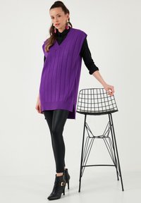 V-neck sleeveless jumper in vibrant purple ribbed knit, paired with black leggings and ankle boots. Wire-styled black chair in background.