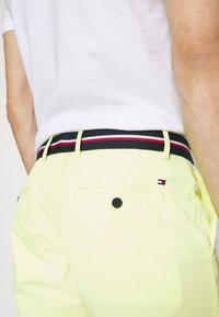 Homme portant un pantalon jaune avec un bouton sur la poche arrière et une ceinture rayée noire, rouge et blanche, associée à une chemise blanche.