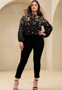 Schwarze Bluse mit floralem Design, transparenten langen Ärmeln, rundem Ausschnitt und Raffung. Kombiniert mit schwarzen, figurbetonten Hosen und schwarzen Riemchen-Heels.
