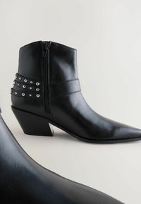Botines de cuero negro con punta afilada, tacón inclinado, cremallera lateral y adornos decorativos en forma de tachuelas plateadas en las correas alrededor del tobillo.