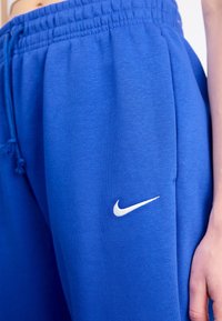 Modré tepláky vyrobené z měkké látky, s elastickým pasem, nastavitelnou šňůrkou a bílým logem Nike na boku.