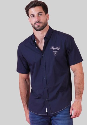 Homme aux cheveux foncés et à la barbe, portant une chemise à manches courtes bleu marine avec le logo "Ruckfield Le Club" et un jean bleu.
