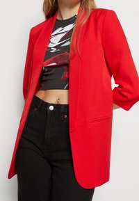 Röd kavaj med sjalkrage, strukturerad form och trekvartsärmar, kombinerad med svarta högmidjade jeans och en grafisk kortärmad topp.