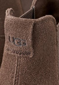 Stivaletto alla caviglia in suede marrone caratterizzato da una texture morbida, cuciture visibili e un piccolo logo UGG sul tallone. Design classico con una forma arrotondata.