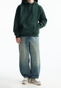 Personne portant un sweat à capuche vert foncé, un jean bleu délavé ample et des baskets gris clair, debout avec une main dans la poche sur fond blanc.