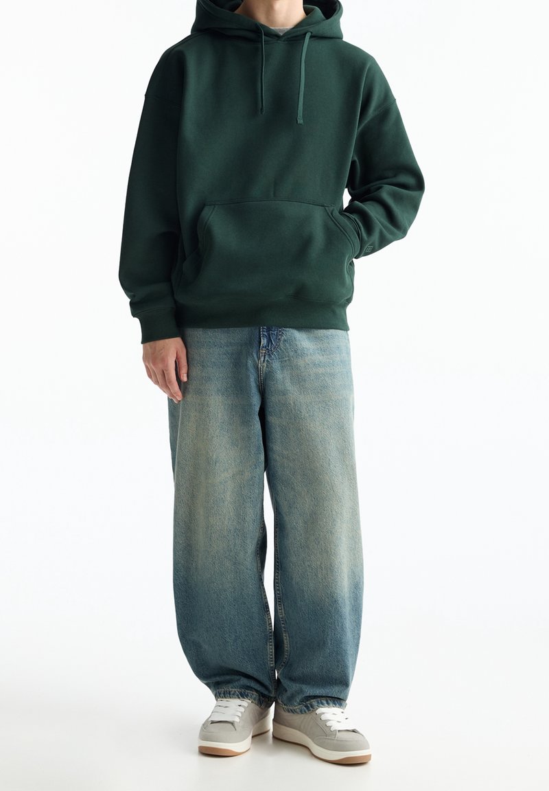 Personne portant un sweat à capuche vert foncé, un jean bleu délavé ample et des baskets gris clair, debout avec une main dans la poche sur fond blanc.