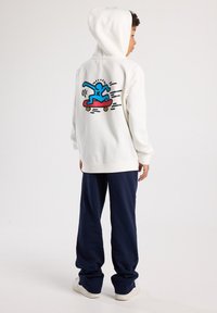 White hoodie with a turquoise skateboard graphic on the back, accompanied by red and black details. Paired with navy pants and white sneakers.